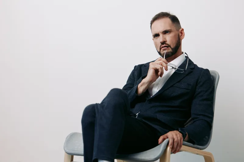 Professional male business figure sits thoughtfully on a chair against an isolated white background, radiating confidence, focus, and calm determination in a modern studio setting.