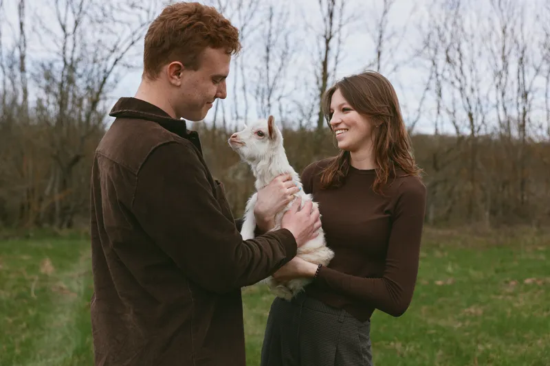 Cheerful couple outdoors, smiling, holding a young goat kid, warm sunlit moment, relaxed posture, rustic setting, candid lifestyle scene, gentle companionship, natural colors, grassy field backdrop