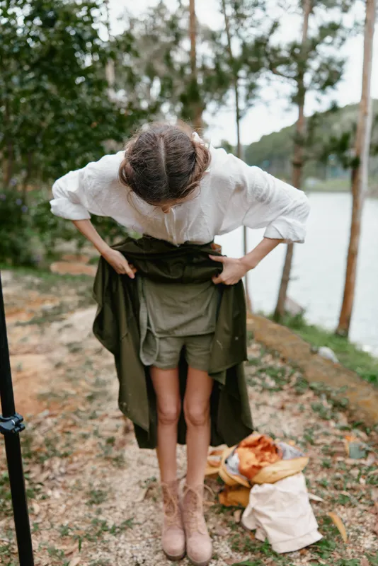 Young woman adjusting her outfit by the river, wearing a casual green dress with a light shirt, showcasing a natural and serene expression surrounded by a lush environment