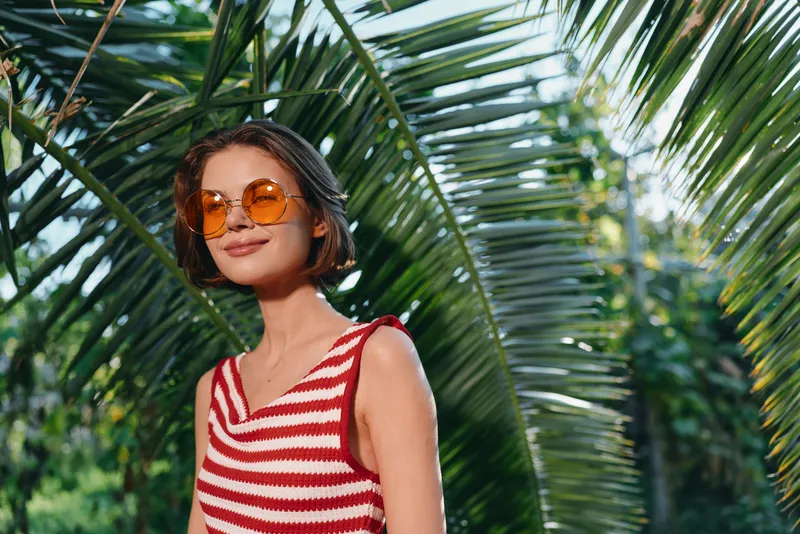 A stylish woman wearing a red and white striped sleeveless top and orange sunglasses stands in a sunny tropical garden with lush palm leaves.