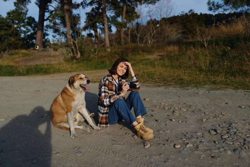 Woman sitting on rocky ground with a dog, enjoying a sunny outdoor moment in nature, casual outfit, relaxed pose and companionship on a lakeside trail.