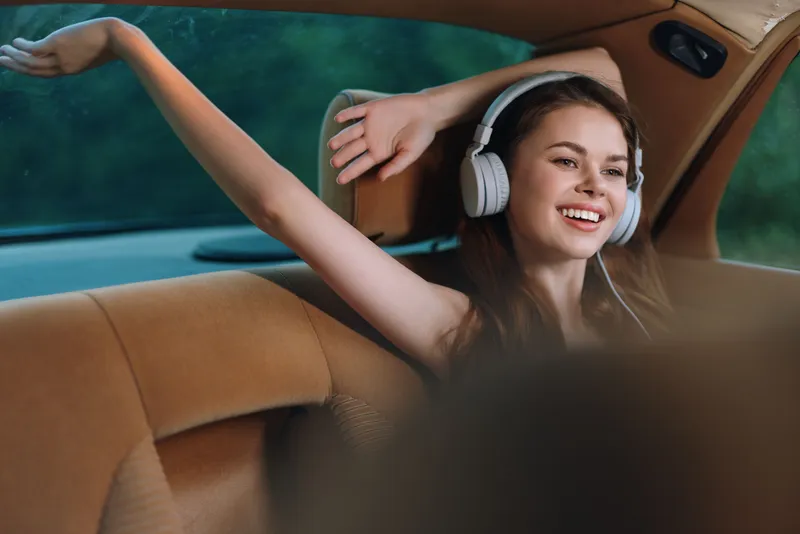 Happy young woman in a car enjoying music with headphones, showcasing joy and relaxation during a road trip, with a warm color palette and natural background.