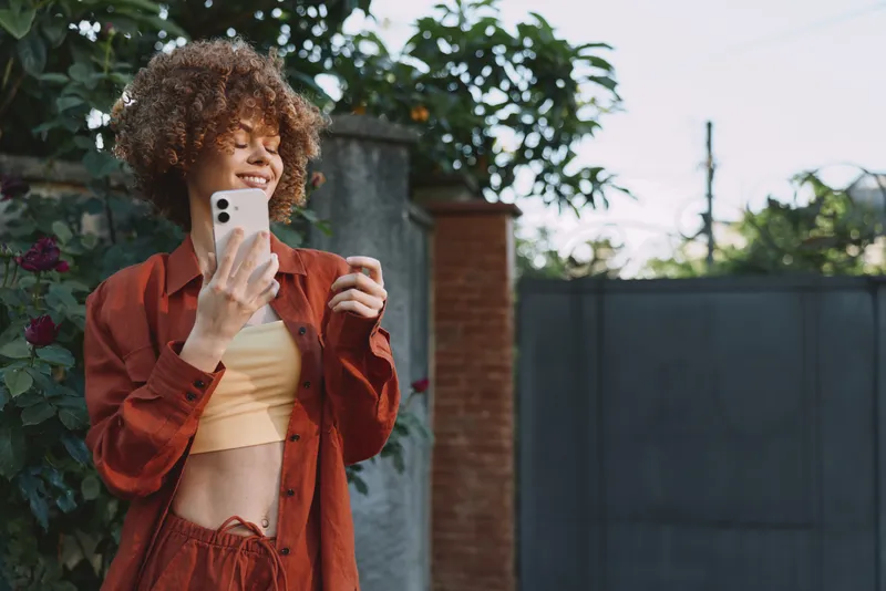 A stylish woman in a red jacket and yellow top takes a casual selfie outdoors, smiling as she captures a candid lifestyle moment in bright daylight, showcasing fashionable style, confidence