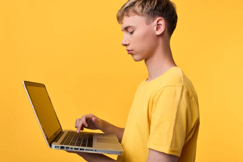 Young man in a yellow shirt uses a laptop, focusing on keyboard and screen in a modern technology scene, casual home studio setup for learning and work