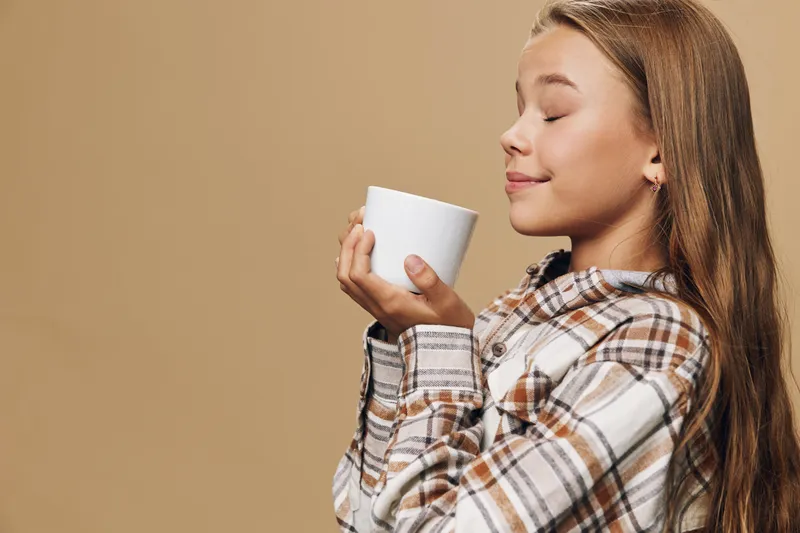 Girl with long hair in plaid shirt holding white cup close to face with closed eyes enjoying warm beverage on beige background for cozy moment and comfort.