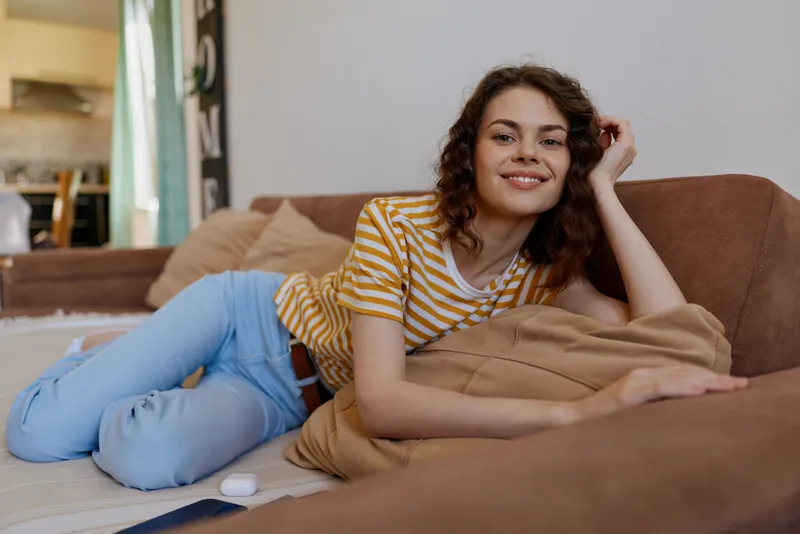 Relaxed woman lounging at home on a couch, casual lifestyle portrait, smiling softly, striped shirt, jeans, cozy living room with natural light and cushions