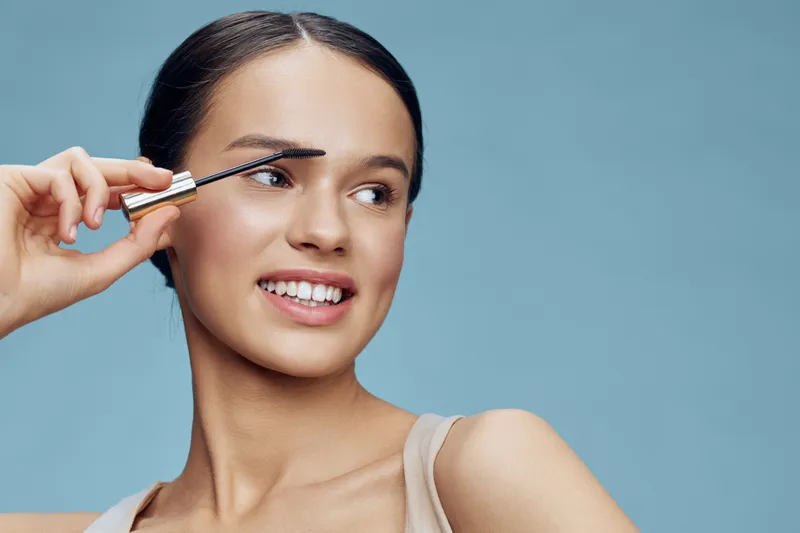 Young woman applying mascara with a smile against a light blue background, showcasing beauty and confidence in a fresh makeup routine
