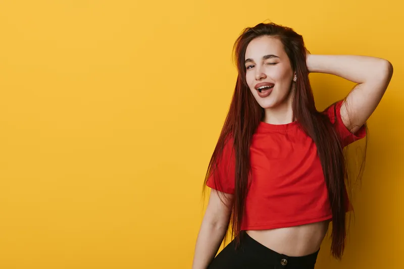Young woman with long hair wearing red crop top posing with eyes closed and hand behind head on yellow background. Casual style, confident smile and relaxed posture in studio.