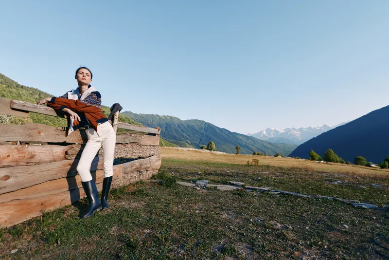 Woman in mountains by wooden fence in countryside field wearing boots and riding clothing, portrait style outdoors leisure with scenic meadow, sky and relaxed fashion pose.