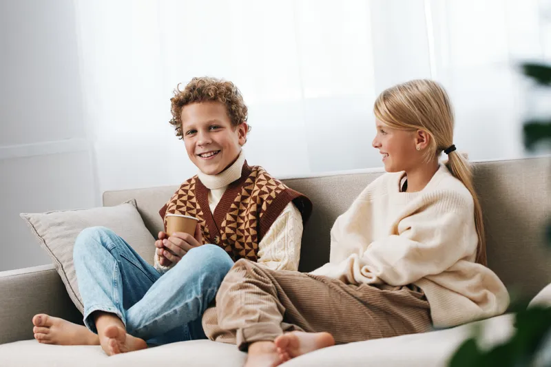 Two cheerful children sit on a sofa, wearing cozy sweaters, sharing a warm moment with a cup of drink, bright living room scene, soft natural light, relaxed mood.