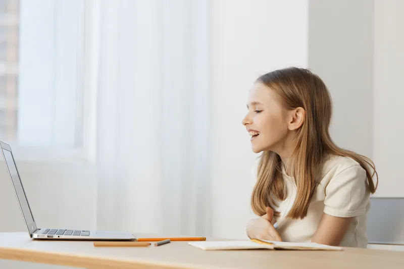 Happy girl engaging with an online lesson at home, wearing a casual white shirt, showcasing joy and excitement in a bright, modern workspace