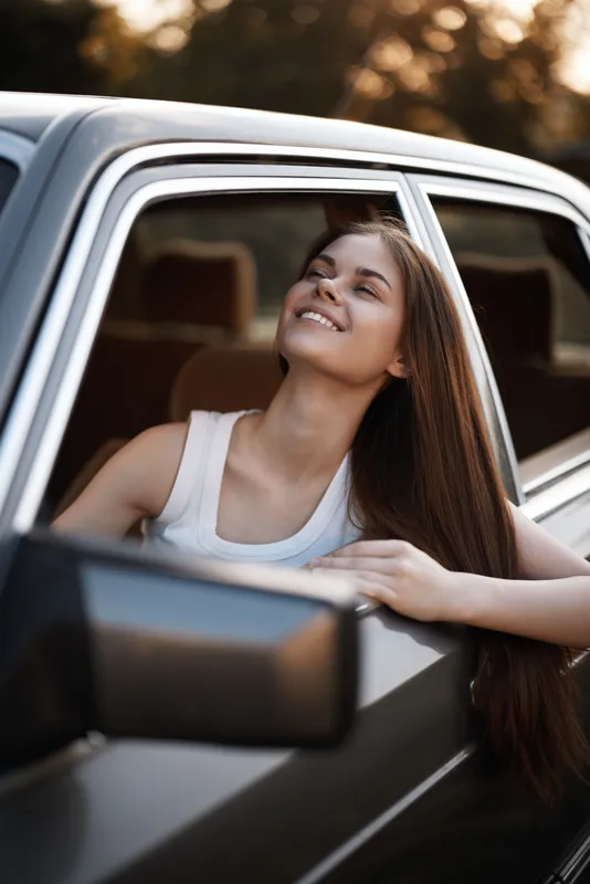 Happy young woman enjoying the breeze while sitting in a car, dressed casually, with a warm sunset background reflecting joy and freedom.