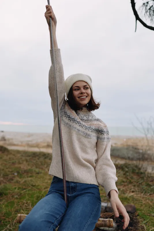 Woman outdoor rope swing on a beach, wearing a cozy sweater and hat, smiling as she enjoys nature and a relaxed casual moment by the sea.