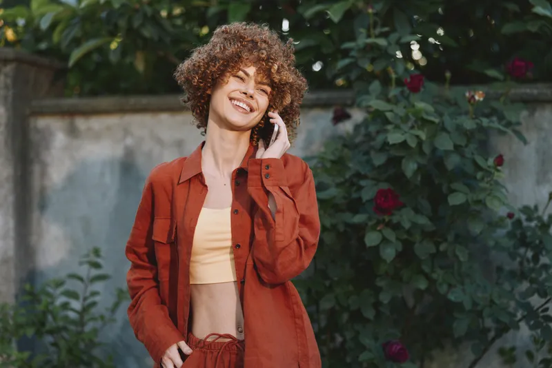 Vibrant woman lifestyle outdoors, smiling while talking on a phone, curly hair, wearing a red jacket, casual crop top, sunlit garden backdrop, natural colors, candid moment