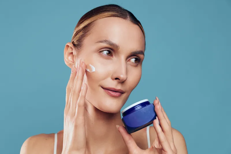 Young woman applies facial cream on clean skin holds blue jar skincare product looks away with soft natural makeup isolated on blue background beauty and health concept.