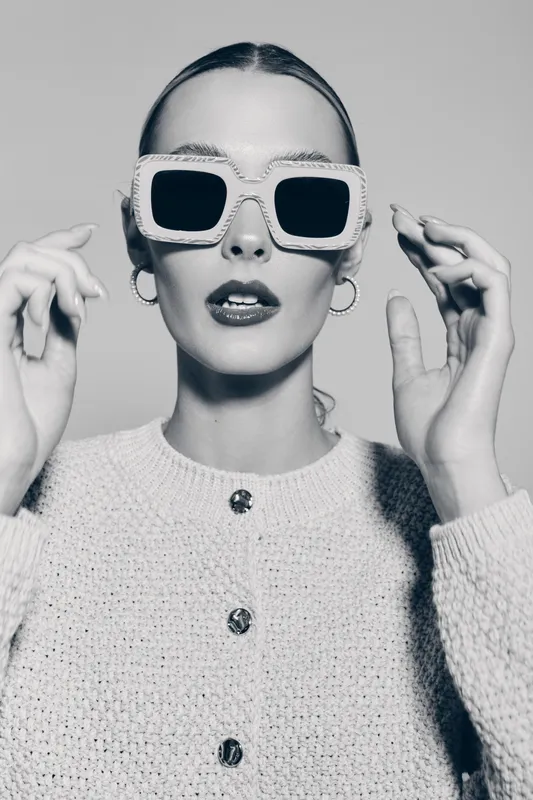 Stylish young Caucasian woman with fair skin wearing large square sunglasses and textured sweater adjusting glasses indoors in black and white portrait.