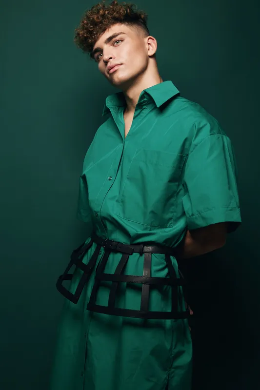 Young fashion model with curly hair wearing green oversized shirt dress with black cage belt styling, posing at studio background for haircut, portrait, style, and beauty concept.