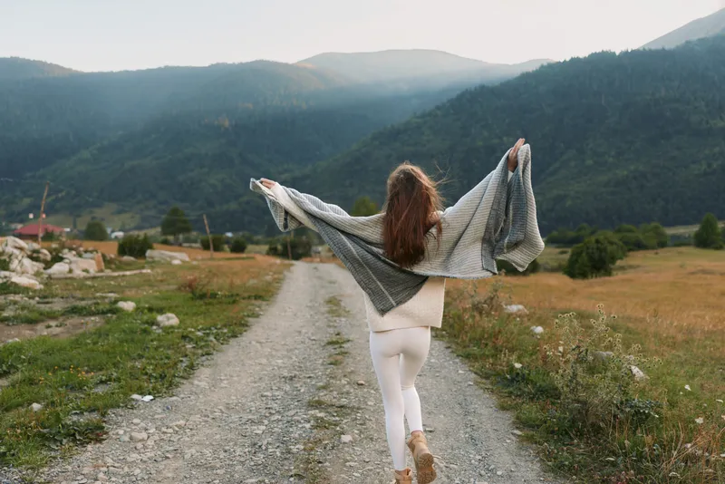 A woman in white leggings and a gray shawl walks on a gravel road in a mountainous rural area with arms raised. The scene shows freedom and lifestyle outdoors.