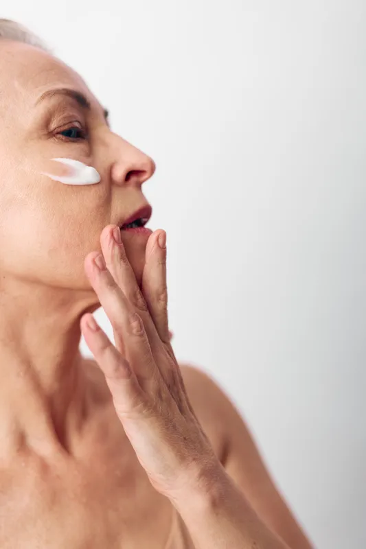Mature woman applies white face cream on cheek with fingers, skincare routine concept. Close-up side view on neutral background highlights natural beauty and self-care for aging skin.