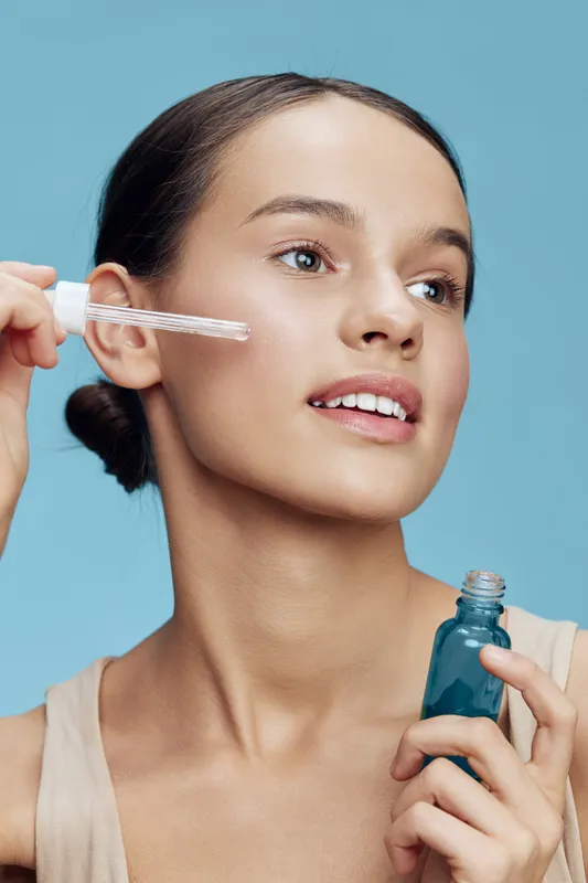 Young female model applying serum on her face with a sapphire bottle in hand, showcasing a radiant and natural skincare routine against a soft blue background