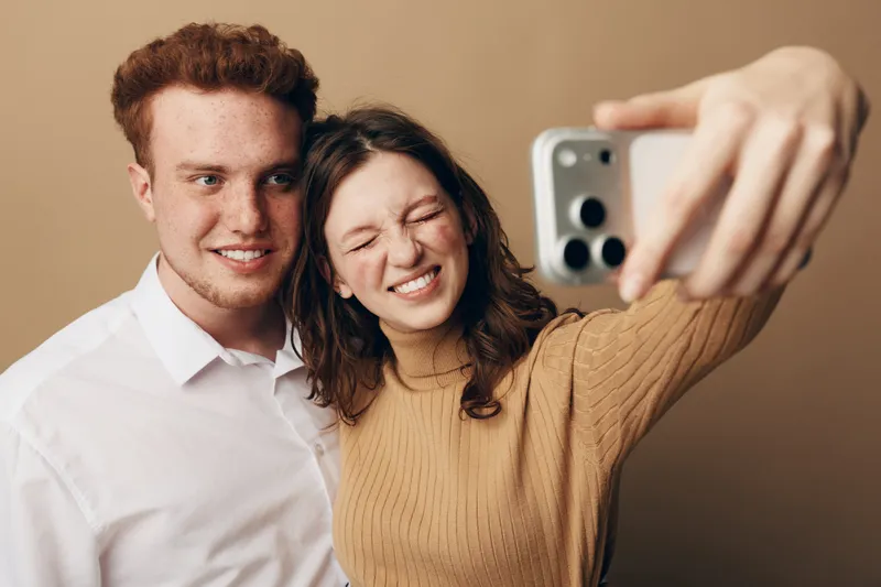 Young couple takes a selfie with a smartphone. The woman smiles with closed eyes while the man looks at the camera. Casual clothing and warm beige background create a cozy mood.
