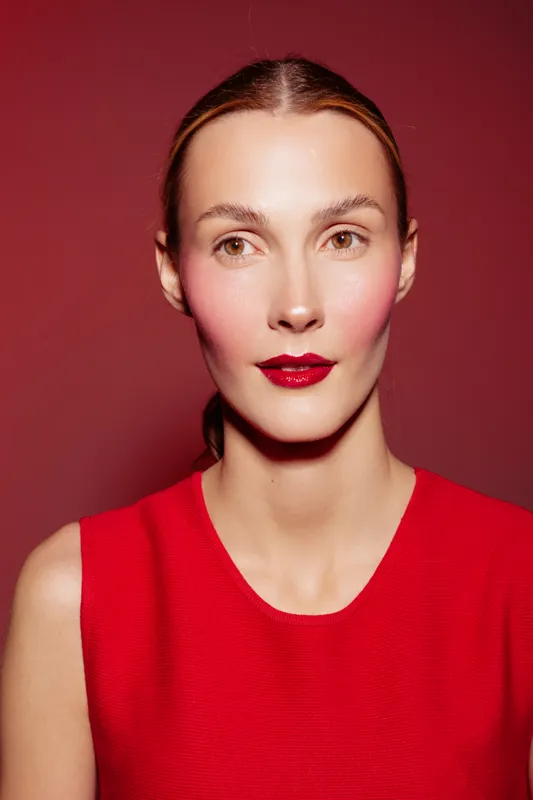 Portrait of a woman with red lipstick and blush wearing a red sleeveless top against a red background. Beauty concept with bold makeup and minimalistic style.