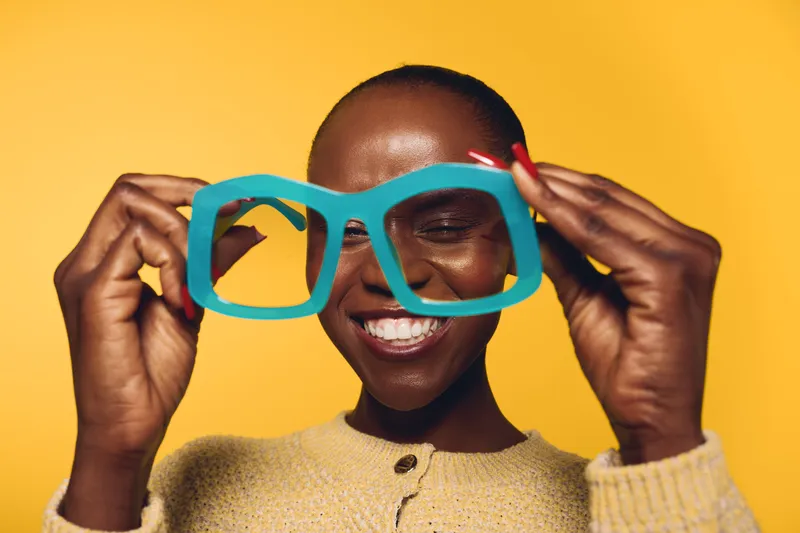 Smiling dark skinned African woman holds oversized blue glasses in front of face on yellow background. Concept of joy, fashion, and playful mood with vibrant colors and happy expression.