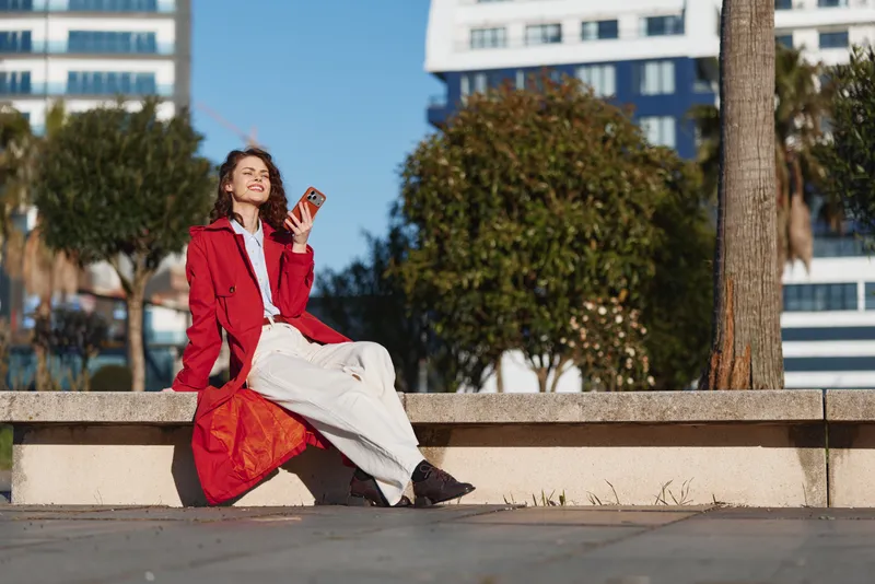 Young woman in red coat sitting on concrete bench outdoors, enjoying sunny day, relaxed posture, modern urban background, casual style outfit