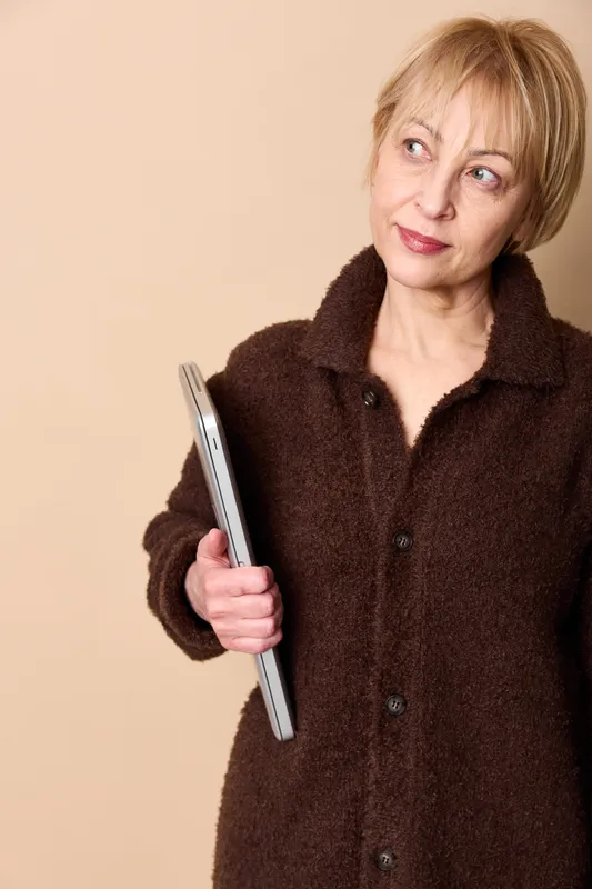 Senior woman portrait, holding smartphone, gentle smile and sideways gaze, warm beige background, cozy coat, technology and communication for older adults