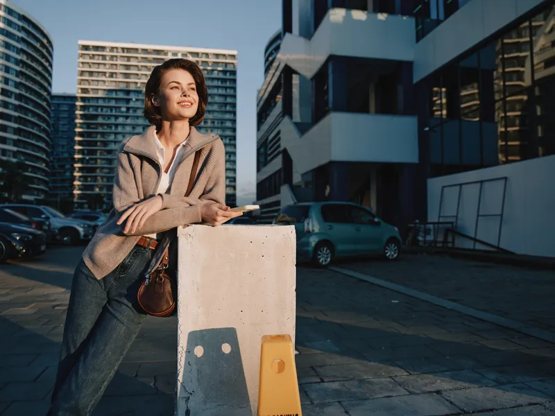 A cheerful young woman stands outdoors beside a modern building, smiling softly while resting on a concrete barrier with a bag, creating a confident urban mood.