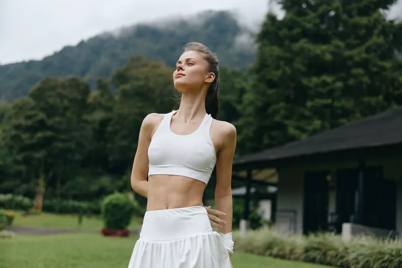 Athletic young woman in white sportswear poses confidently outdoors, showcasing fitness and vitality against a beautiful natural backdrop with greenery and mountains