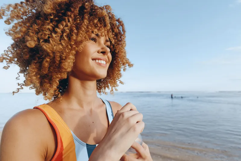 A smiling woman on a sunny beach poses in colorful swimwear, holding a water bottle. Bright sky, calm sea behind. Concept: wellness, outdoor living, summer mindset and self-care.