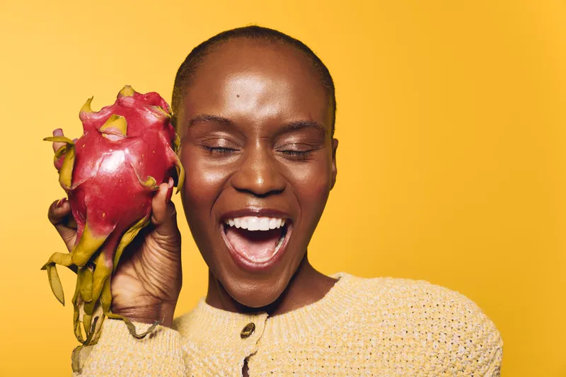 Smiling dark skinned African woman holds fresh dragon fruit near face with eyes closed. Yellow background and beige sweater create vibrant and joyful studio portrait. Concept of health and happiness.
