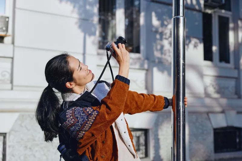 Binoculars, woman, outdoor observation of urban landscape with building and daylight, exploration and curiosity as she scans the street from a pole side position for scouting and research in motion