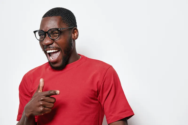 African American man with dark skin wearing glasses and red t shirt smiling and pointing finger on white background. Happy male portrait with casual style and positive expression.