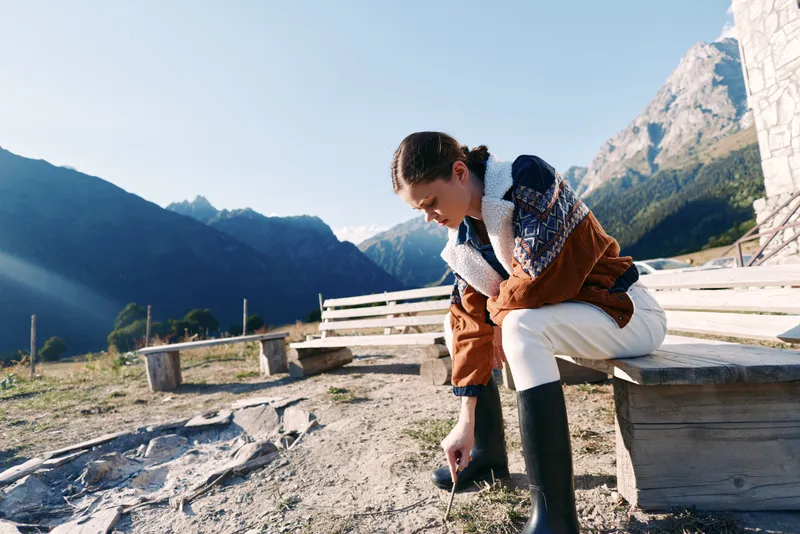 Woman sits on wooden bench in mountain landscape, wearing sweater and waterproof boots while tying laces before hiking. Outdoors nature scene with clear sky and alpine peaks.