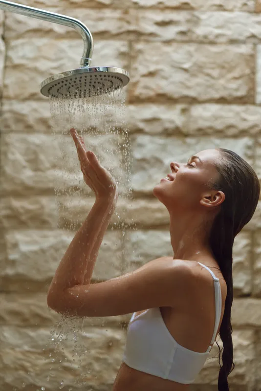 Young woman enjoying warm shower water with eyes closed, wet hair, and relaxed expression against rustic stone wall background, wellness and self-care moment.