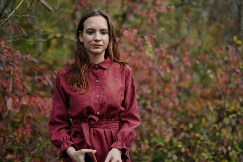 A young woman stands in autumn woodland wearing a simple red dress, captured in a candid, authentic portrait conveying genuine emotion and natural presence among colorful foliage and soft light.