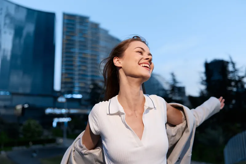 happy woman enjoying freedom outdoors in casual white shirt with open arms against urban blue sky background smiling joyful cheerful