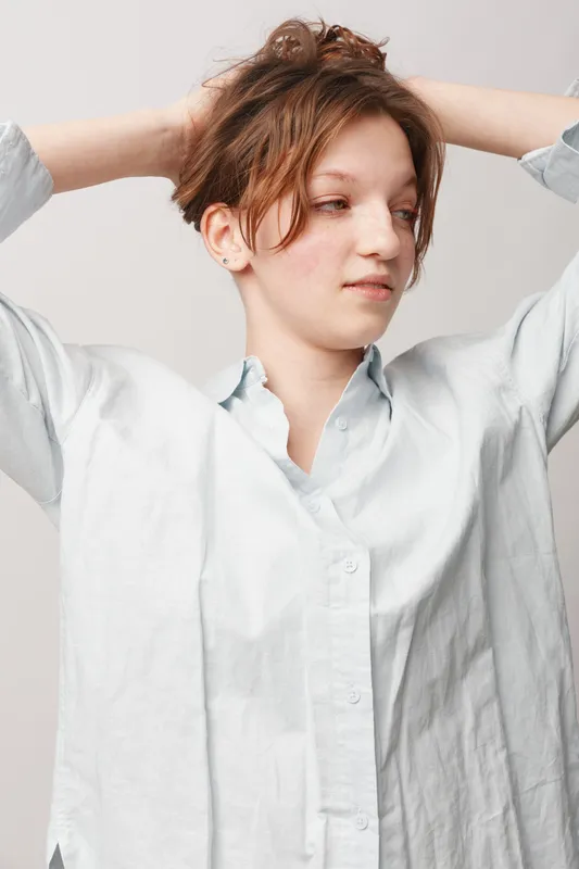 Young woman with short brown hair wearing white shirt looks away with hands behind head. Casual style, natural light, relaxed mood, isolated on light background.