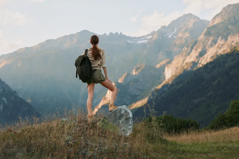 Woman hiker with backpack stands on a rock in a mountain meadow, enjoying nature and outdoor scenery during daytime, solo travel vibe.