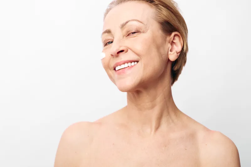 Smiling middle aged woman with short hair applies white face cream on cheek. Close up on white background. Concept of skincare, beauty, anti aging, wellness, and self care for mature skin.