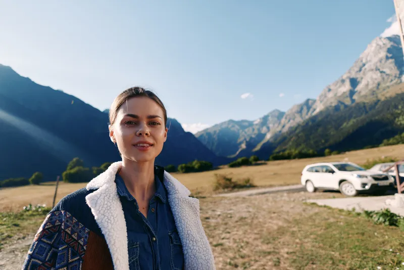 Woman portrait in countryside with mountains and car in background, travel and outdoors scene with nature landscape, meadow road and clear sky for adventure and leisure.