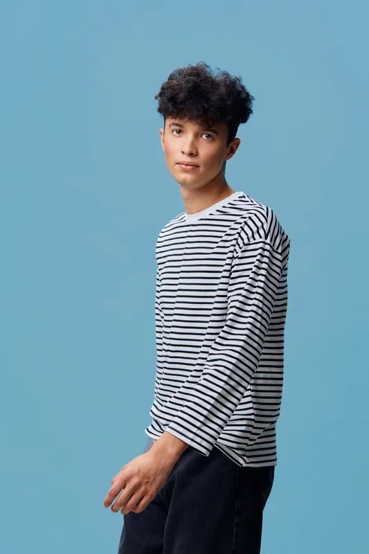 Young man with curly hair wearing a striped shirt standing against a blue background, looking thoughtful and casual in a minimalist studio setting.