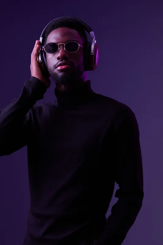 Stylish young Black man listening to music with headphones, wearing sunglasses, against a vibrant purple background, expressing confidence and relaxation