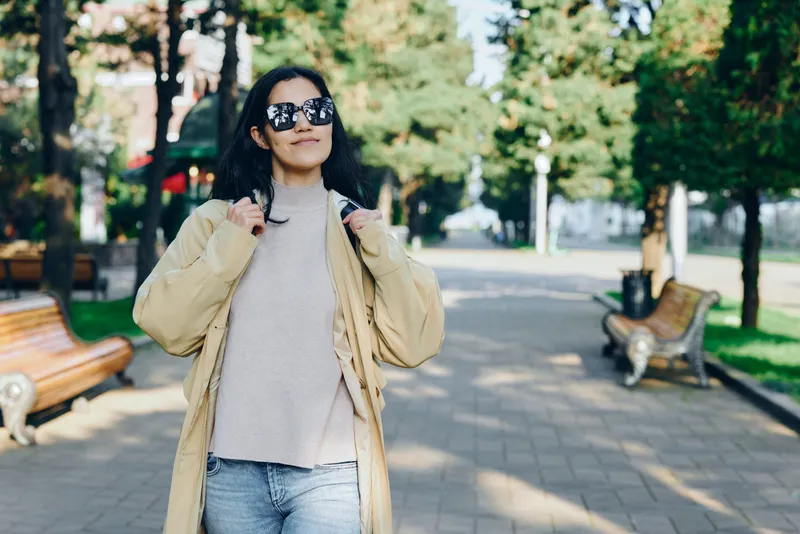 Woman strolling in a sunlit park wearing a beige coat and knit sweater, sunglasses on, casual chic styling in an urban street scene, lifestyle oriented outdoor photography that captures confident,