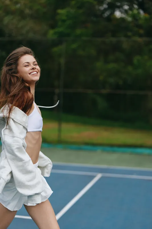 Happy young woman enjoying summer on the tennis court, wearing a white crop top and shorts, showcasing a carefree spirit in a lush green background