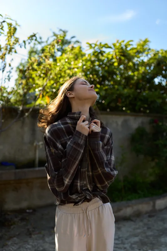 A confident woman stands outdoors in soft daylight, looking up with a gentle smile. A plaid shirt and light trousers create casual, relaxed style in a sunlit garden