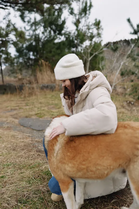 Woman in a warm jacket and beanie outdoors with a large dog, kneeling on grass, sharing a gentle moment of companionship in a cold countryside setting.