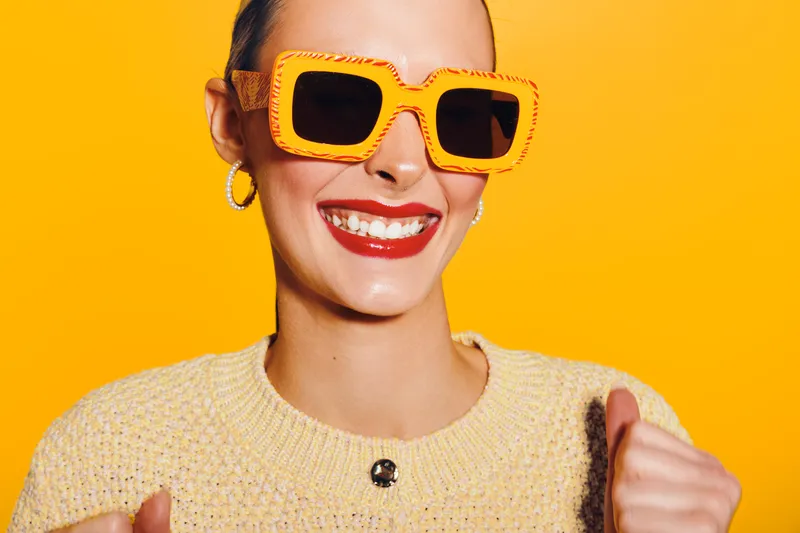 Smiling woman with bright lipstick wearing yellow sunglasses and knitted sweater on yellow background. Concept of happiness, fashion, summer style, and positive mood.